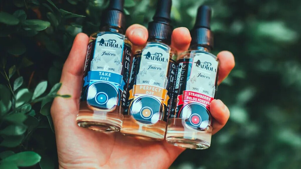 A close-up, eye-level shot of a hand holding three bottles of Radiola Juices vape liquid in a lush, green outdoor setting. The bottles are labeled "Take Five," "Perfect Day," and "Strawberry Fields Forever," each featuring a unique color scheme and a design resembling a vinyl record. The hand is holding the bottles horizontally against a backdrop of blurry green leaves, with natural light highlighting the liquid inside the clear bottles.