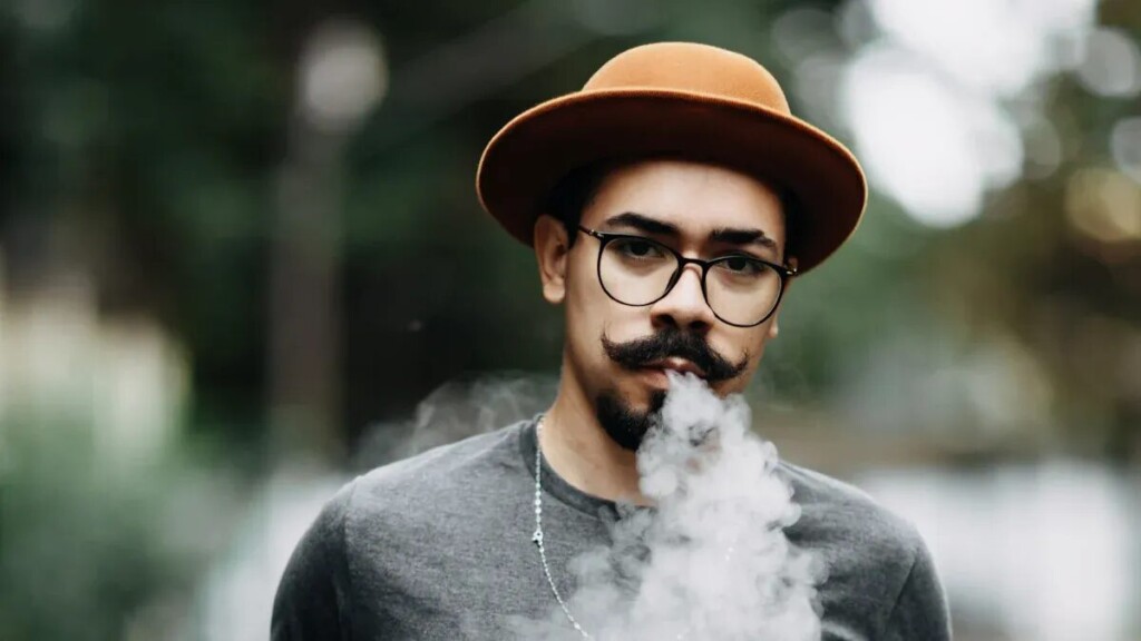 A man with a mustache and goatee, wearing a gray t-shirt, glasses, and a fedora, exhales a large plume of vapor. The background is a blurry outdoor setting with greenery.