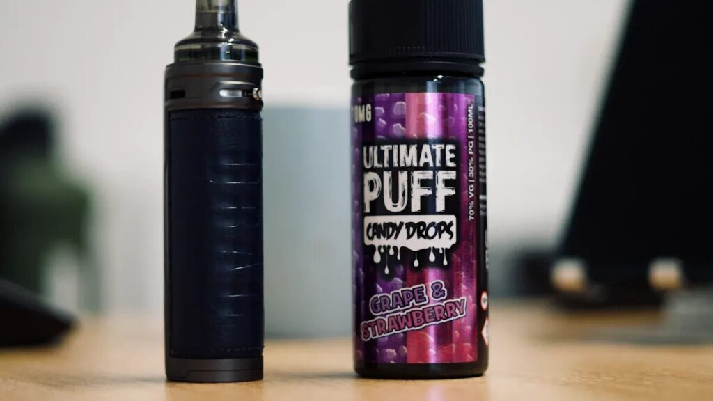 A close-up shot of a dark-colored vape device with a leather-like texture, standing upright next to a bottle of "Ultimate Puff Candy Drops" e-liquid in the flavor "Grape & Strawberry." The bottle is purple and pink with a dripping candy design. The items are on a wooden surface with a blurred background.