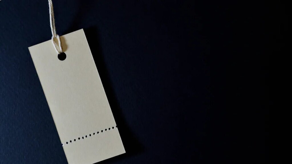  A close-up shot of a plain, blank rectangular tag with a perforated line near the bottom. The tag is made of light-colored cardstock and hangs from a string against a dark blue or black background.