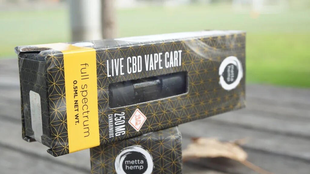 An eye-level, close-up shot of a black and gold box for a "LIVE CBD VAPE CART" from the brand "metta hemp," sitting on a wooden surface outdoors. The box features a complex pattern of geometric shapes. The text on the packaging indicates it contains "full spectrum" CBD, "0.5ML NET WT." and "250 MG CANNABINOIDS." A small, red warning icon with a skull and crossbones is also visible. The background is a blurry outdoor scene with green grass and a tree.